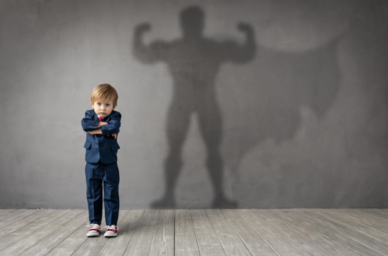 Photo d'un petit garçon qui croise les bras l'air en colère et sur le mur derrière lui est projeté l'ombre d'un homme musclé qui montre ses biceps musclés pour illustrer l'intérêt de la Fight thérapie poiur apprendre aux enfants à se protéger et se défendre par exemple en cas de harcèlement scolaire